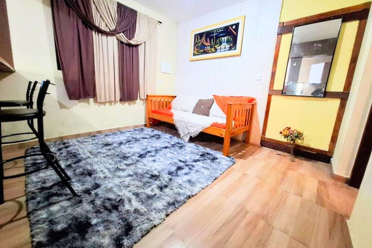 Una habitación con un tapete azul y esponjoso, un sofá de madera con cojines naranjas, una silla de madera, una mesita y un cuadro en la pared.