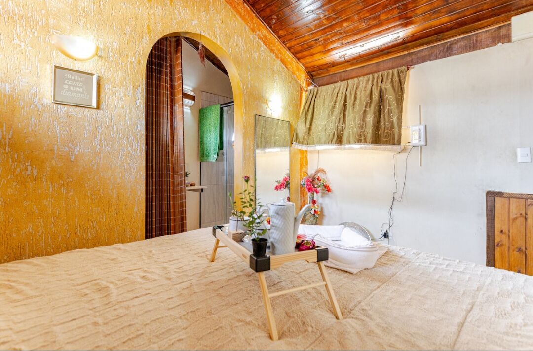 Una habitación con techo de madera, una cama con colcha blanca, una pequeña mesa con un jarrón y flores, y un espejo en la pared.