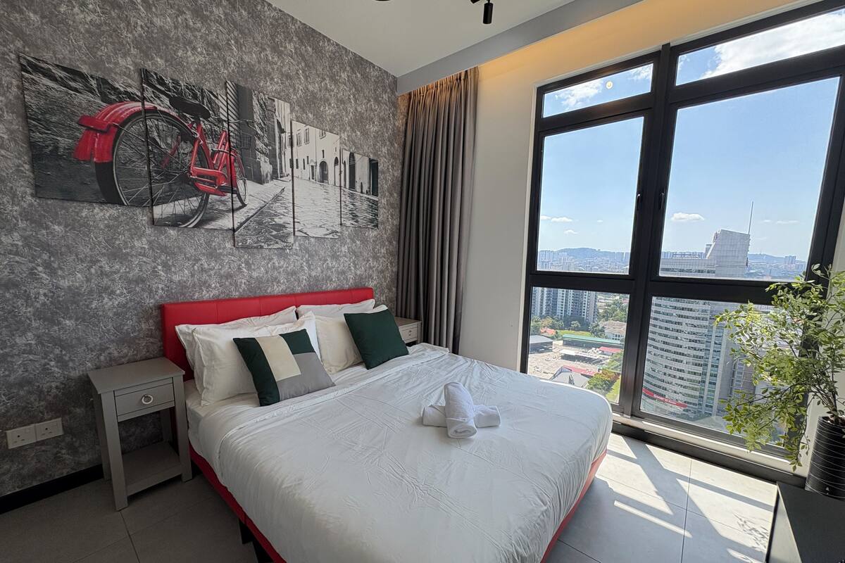 A modern hotel room with a large window, a bed with white linens, a nightstand, and a cityscape view.