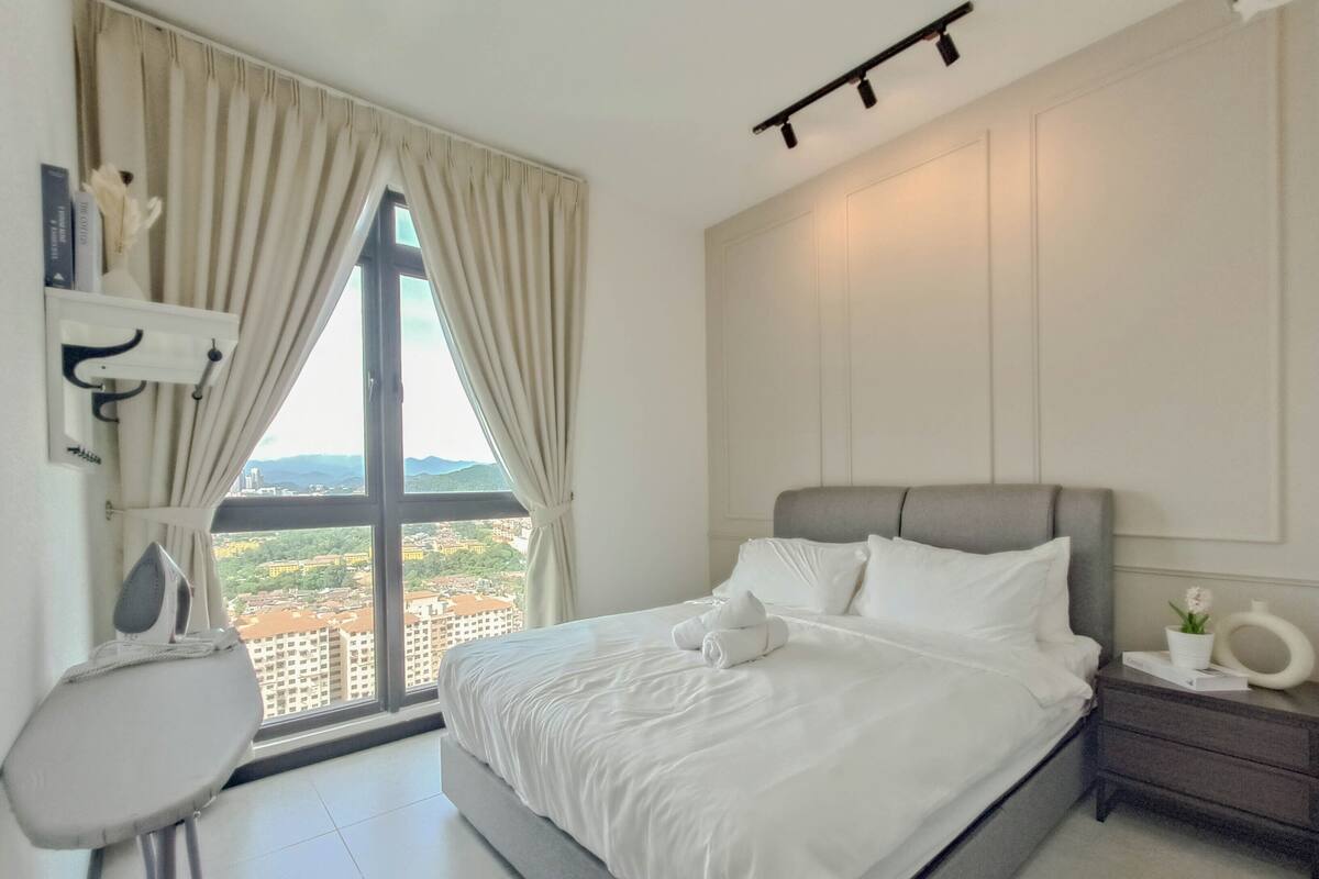 A modern hotel room with a large bed, a desk, and a view of a cityscape through a large window.