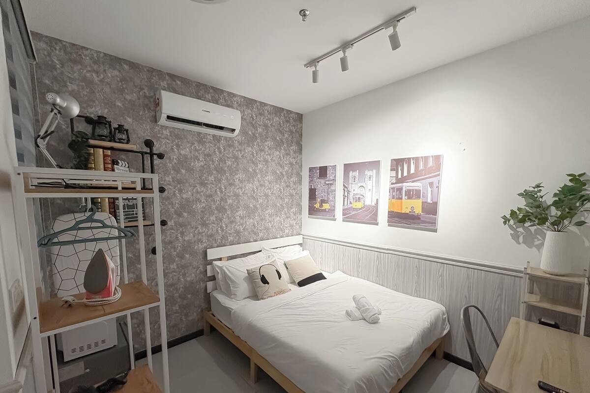 A neatly arranged bedroom with a bed, a shelf with books and a lamp, and a wall-mounted air conditioner.