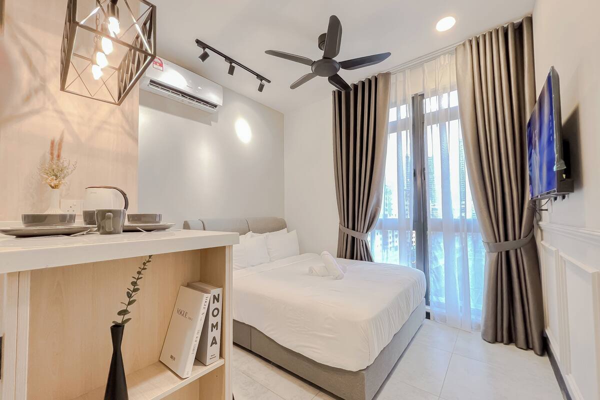 A modern hotel room with a bed, a vanity with a mirror, a ceiling fan, and a large window with curtains.
