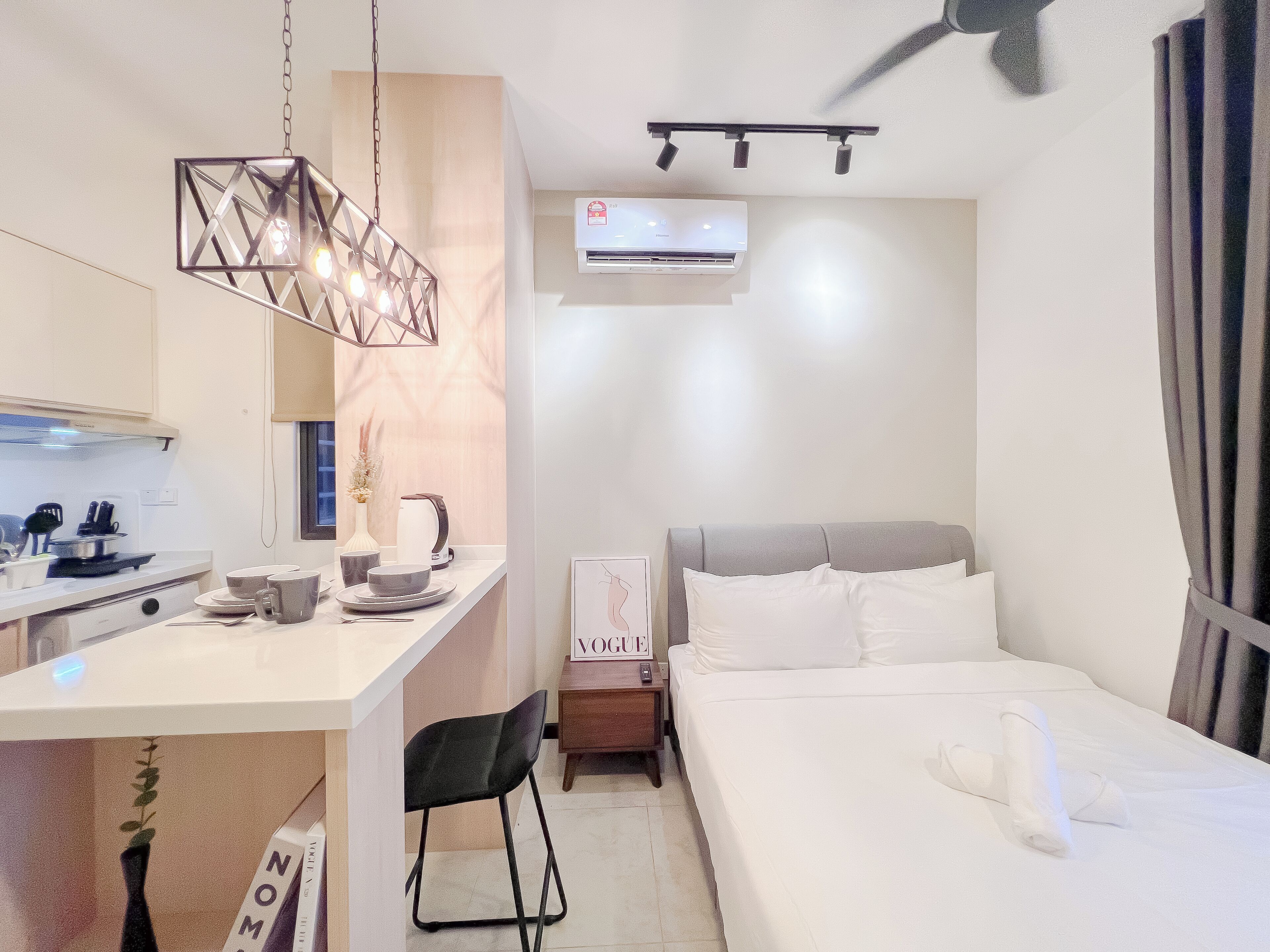A compact hotel room with a kitchenette, a bed, a bedside table, a chair, and a ceiling fan.