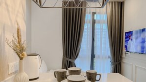 Un comedor moderno con una mesa blanca dispuesta para cuatro, un florero con plantas secas y un televisor mostrando a una persona.