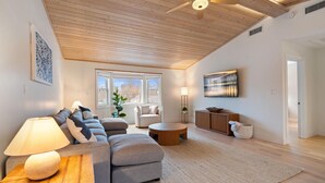 A modern living room with a high ceiling, wooden beams, a sofa set, a coffee table, a floor lamp, a TV, and a large window.