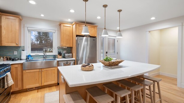 A modern kitchen with wooden cabinets, stainless steel appliances, a large island with stools, pendant lighting, and a view through the window.