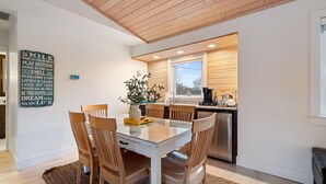 A modern kitchen with wooden ceiling beams, a dining area with a glass table and wooden chairs, and a kitchen island with a coffee machine.