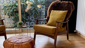 Une pièce chaleureuse avec un plancher en bois, un pouf en cuir, une chaise en bois recouverte d’une peau de fourrure et une table en verre sur laquelle est posée une plante.
