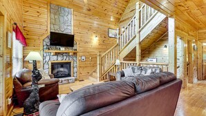 A log cabin interior with a stone fireplace, leather sofa, and a staircase. - Family Cabin with Hot Tub, Theater, Game Room, Wifi, & Sauna (Sevierville)