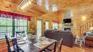 A wooden cabin interior with a dining table set, a sofa, a fireplace, and a TV mounted on the wall. - Family Cabin with Hot Tub, Theater, Game Room, Wifi, & Sauna (Sevierville)