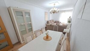 A dining area with a white table, chairs, and a chandelier. There is a sofa and a cabinet in the background.