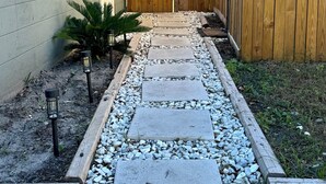 En trädgårdstig som leder fram till ett trästaket, med ett hus på ena sidan och en pergola på den andra.