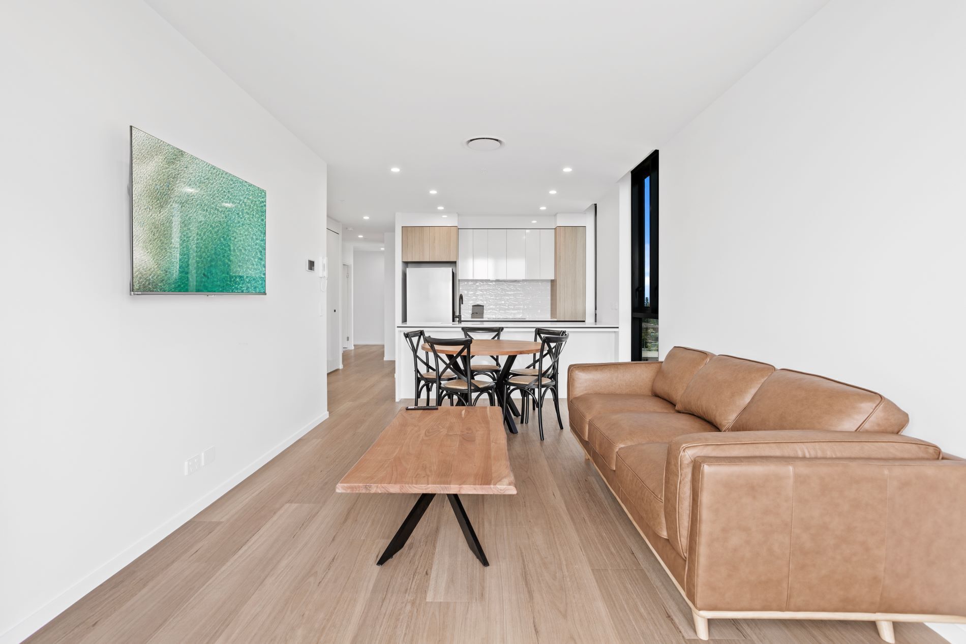 A modern living room with a brown leather sofa, a wooden dining table with chairs, and a kitchen in the background.