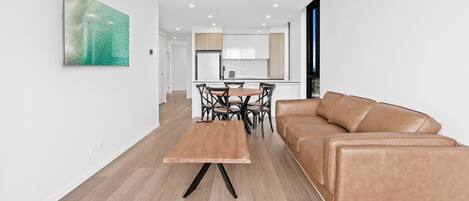 A modern living room with a brown leather sofa, a wooden dining table with chairs, and a kitchen in the background.