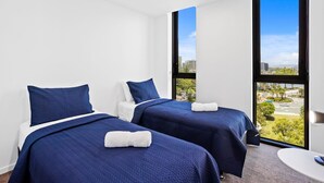 Quarto de hotel com duas camas, cada uma com colcha e almofadas azuis, uma mesa de cabeceira branca e uma janela grande com vista para a cidade.