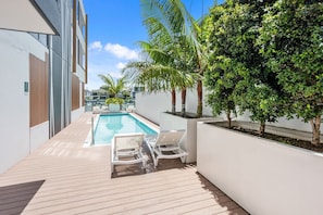 Zona de piscina exterior moderna con terraza de madera, sillas reclinables blancas y vista al mar.