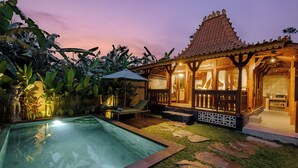 A poolside area with a lounge chair and umbrella, a small pavilion with a tiled roof, and a stone pathway leading to a building with a tiled roof.