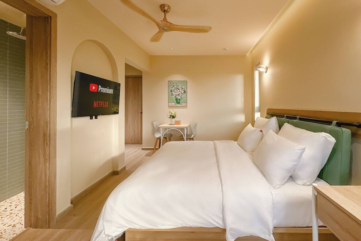 Habitación de hotel con cama, televisión montada en la pared, una mesita con sillas y un ventilador de techo.
