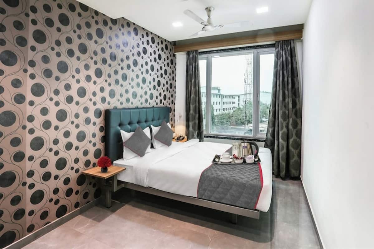A modern hotel room with a polka dot wallpaper, a large bed with a dark headboard, a nightstand with a lamp, and a view of the city through a large window.