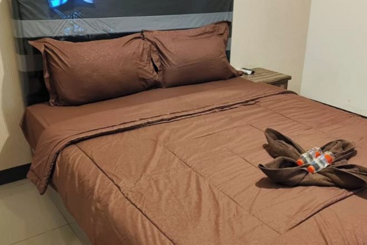 Un lit recouvert d’une courtepointe marron et agrémenté d’oreillers, une table de chevet en bois avec une lampe, et une fenêtre donnant sur le ciel.