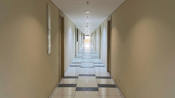A long, narrow hotel corridor with a checkered floor pattern, doors on both sides, and a ceiling with recessed lighting.
