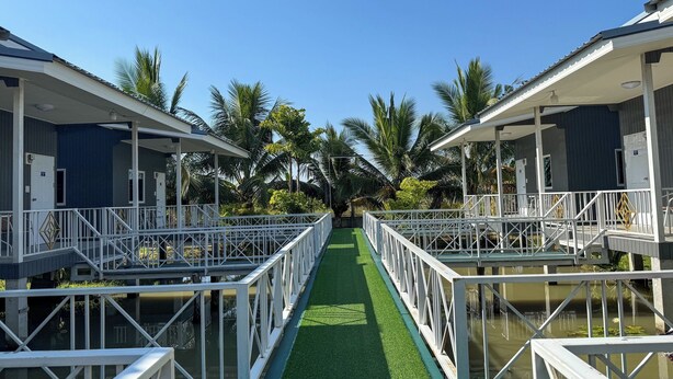 A row of blue and white cabins with white railings, connected by a green pathway over water, surrounded by palm trees. - Jira Hotel (Lampang)