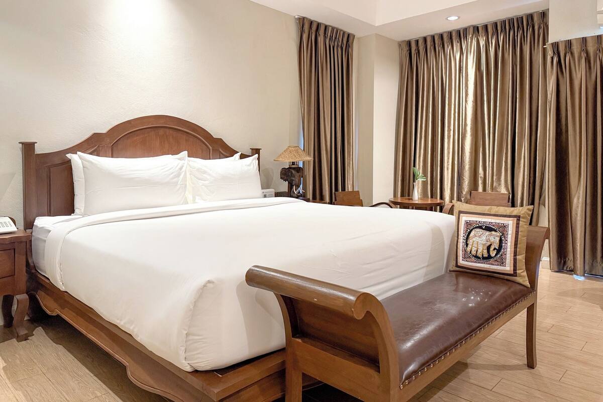 A hotel room with a large bed, wooden headboard, a bedside table with a lamp, a small round table with a plant, and a bench with a decorative pillow.