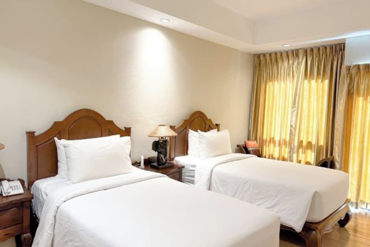 Une chambre d’hôtel avec deux lits simples, des têtes de lit en bois, des tables de chevet avec des lampes et une grande fenêtre avec des rideaux.