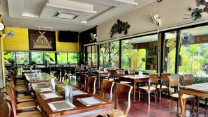 A restaurant with wooden tables and chairs, large windows with a view of greenery, and ceiling fans. - Kokhaburi Hotel (Ko Kha)