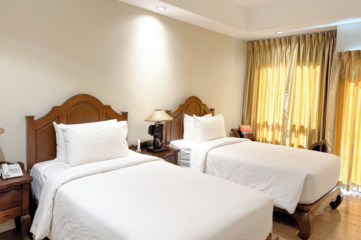 A hotel room with two single beds, wooden headboards, a bedside table with a lamp, a telephone, and a curtain with a golden pattern.
