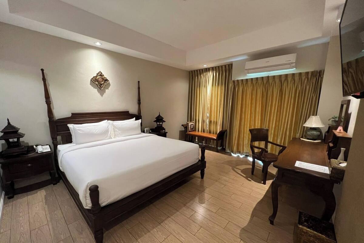 A hotel room with a large bed, a desk with a chair, an air conditioning unit, and a traditional decorative piece on the wall.