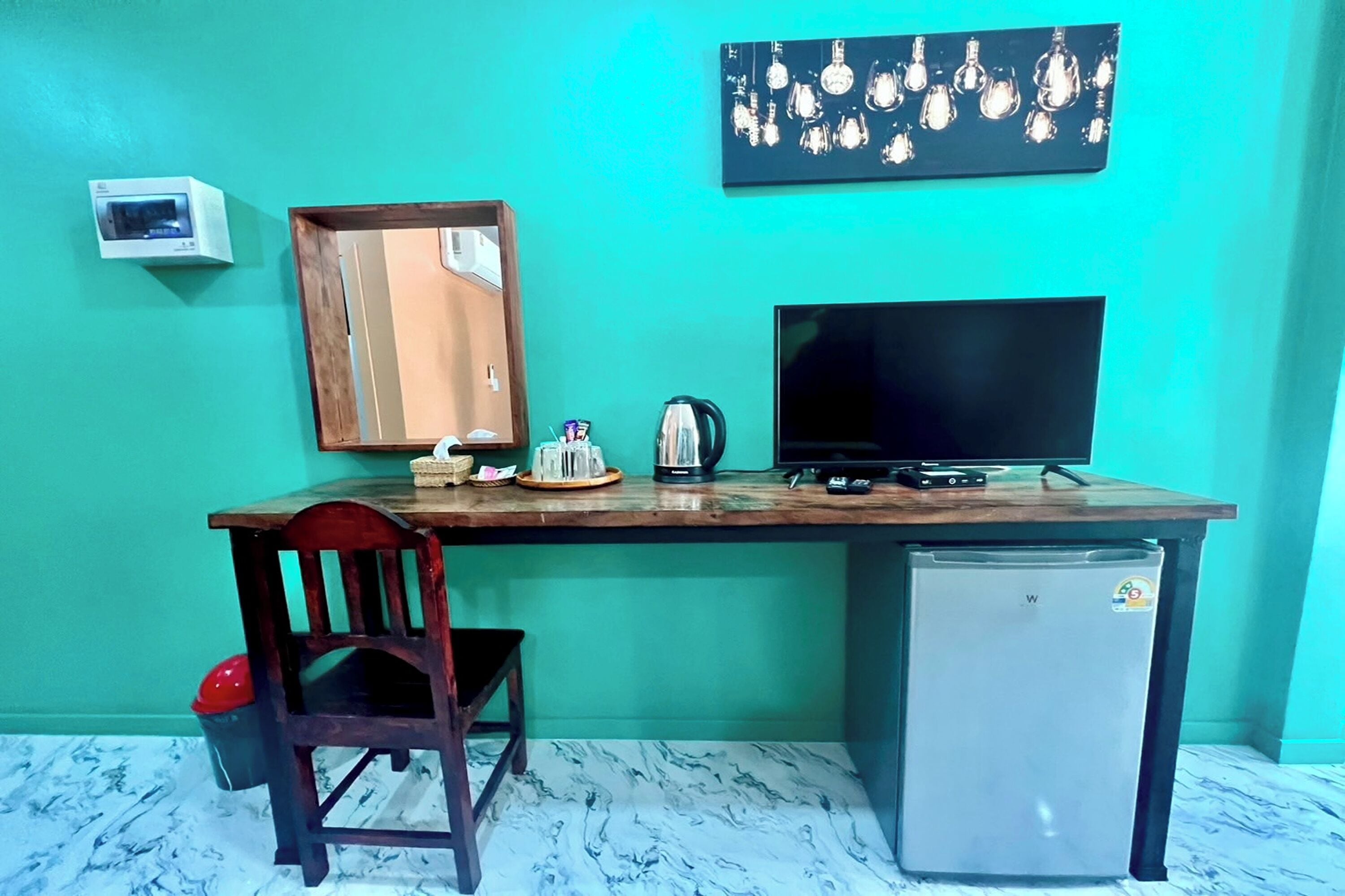A compact room with a teal wall, a wooden desk, a small refrigerator, a television, a chair, and a wall-mounted heater.