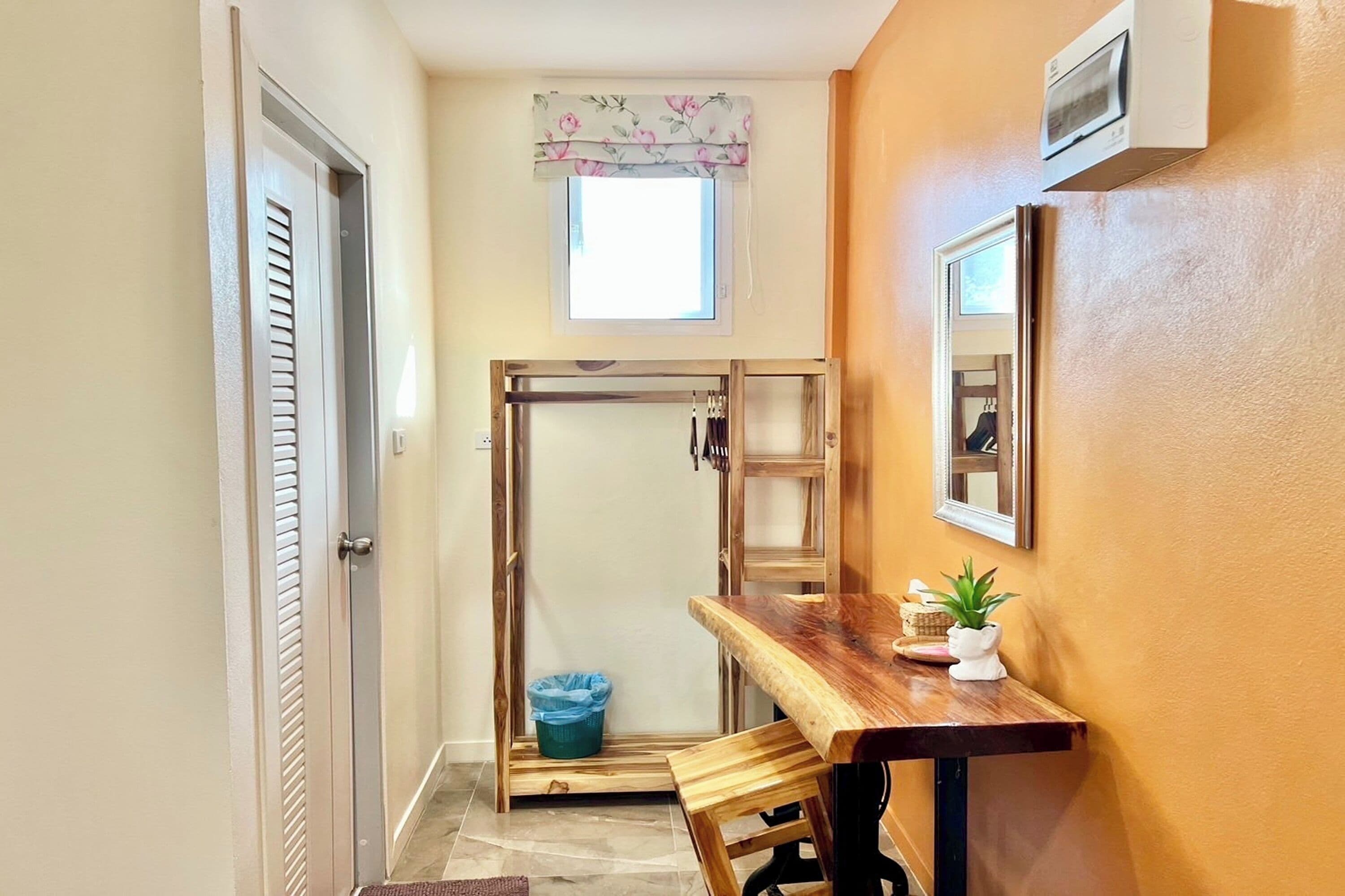 Un couloir étroit avec un banc en bois, une petite table, une fenêtre ornée d’une lambrequin à motifs floraux et un miroir accroché au mur.