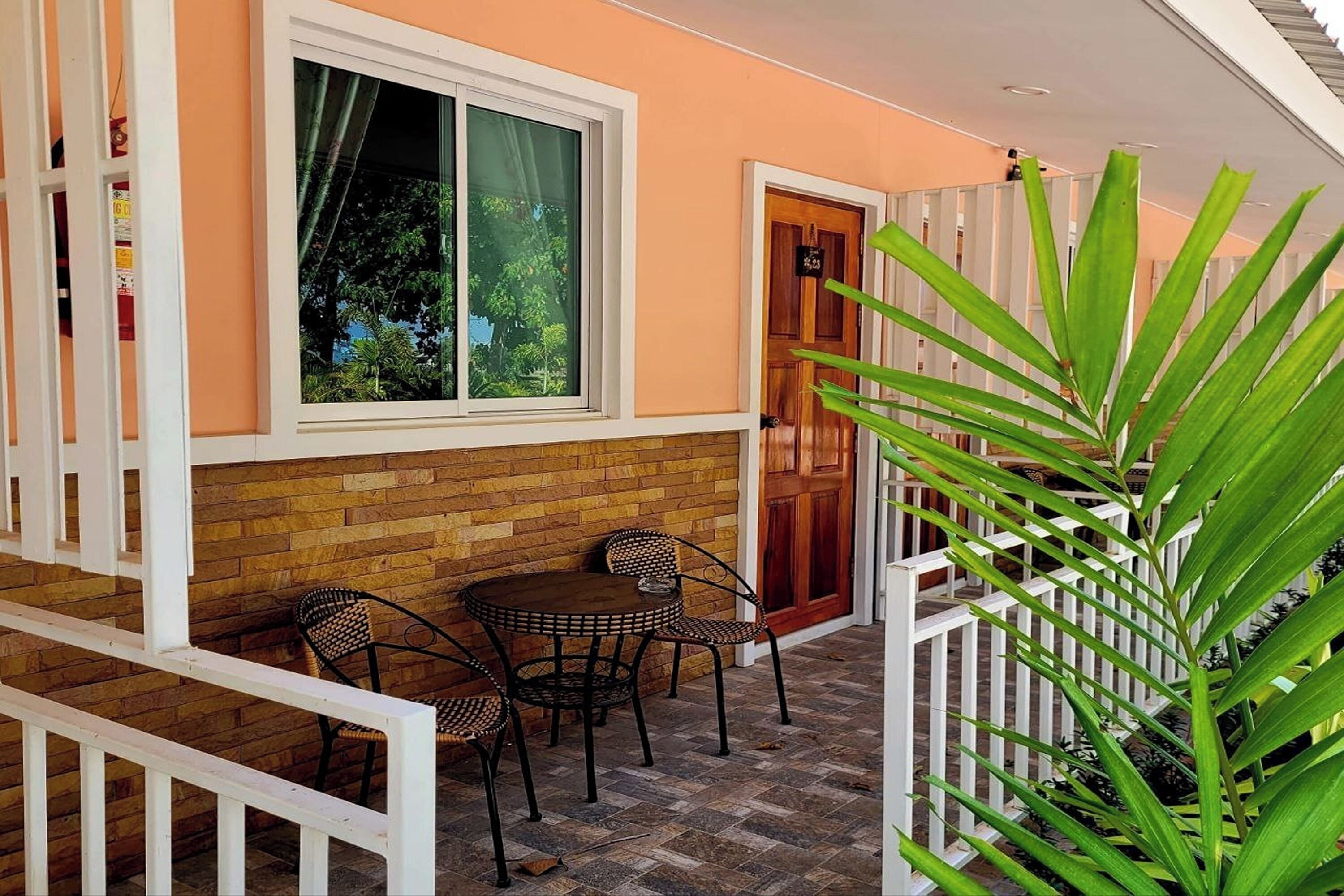Un patio con un muro de ladrillo, una ventana y una puerta. Hay dos sillas de mimbre y una pequeña mesa redonda.