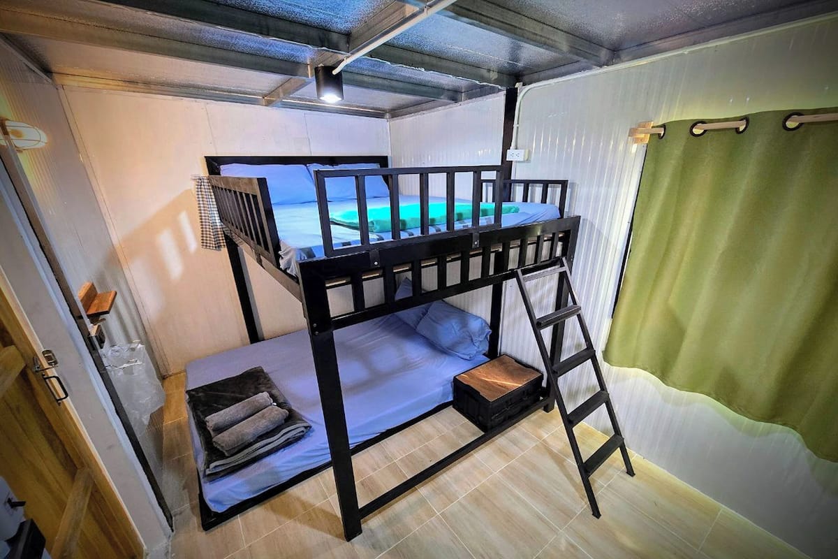 A bunk bed room with a view of a turquoise sea through a window, a ladder, and a green curtain.