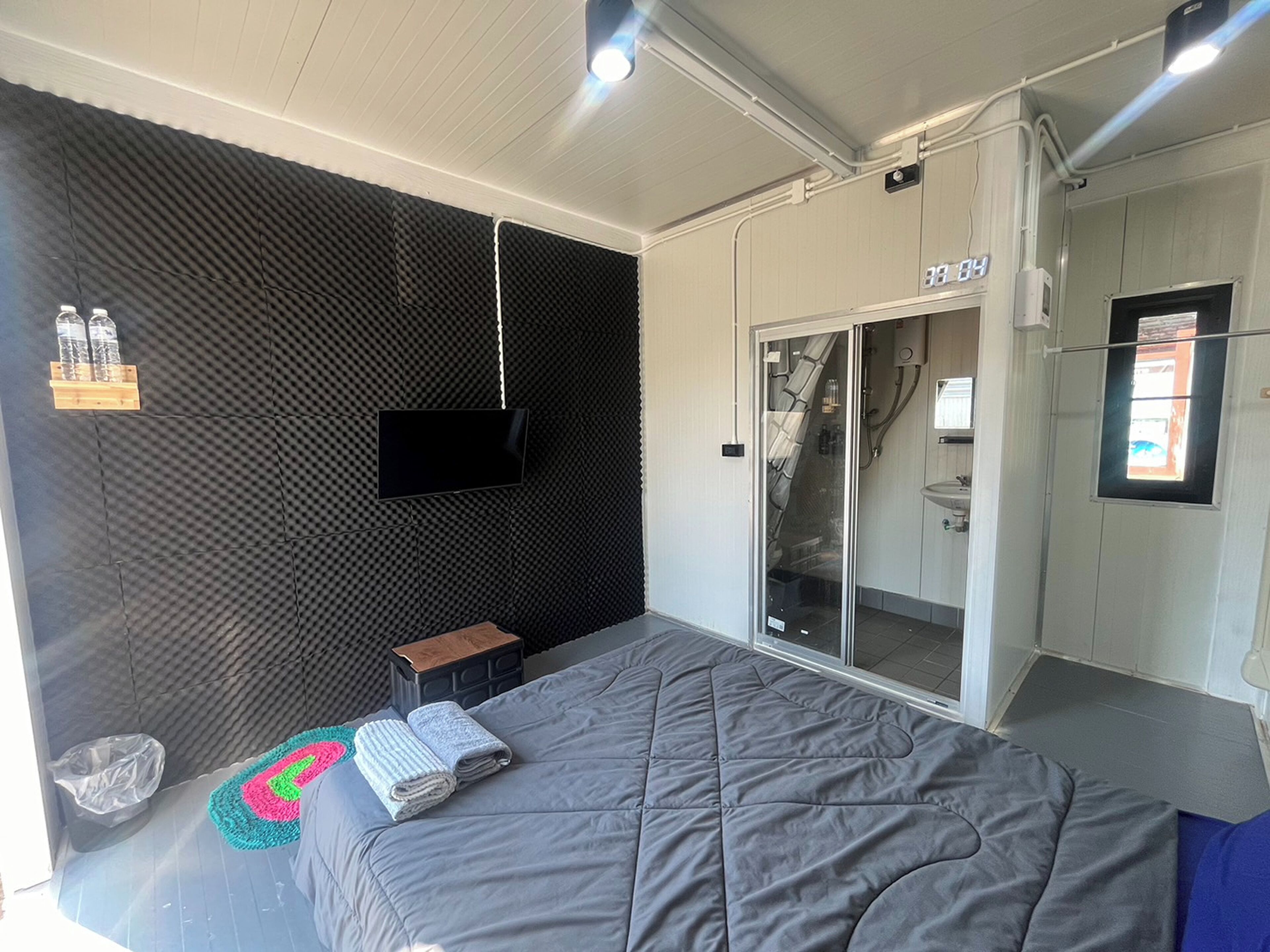 A modern bedroom with a grey bed, a flat-screen TV mounted on a black acoustic panel, a small wooden stool, and a bathroom with a glass door.