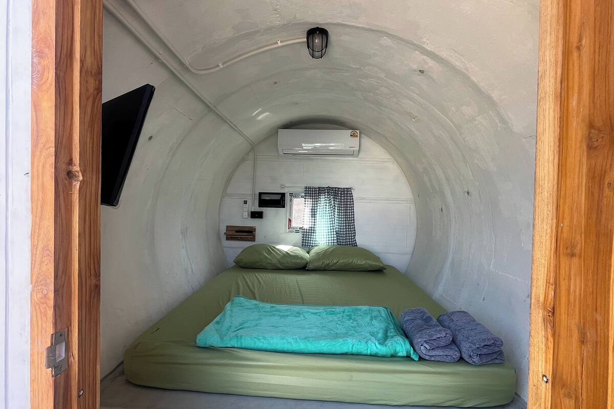 A small, enclosed room with a single bed, a window with checkered curtains, an air conditioning unit, and a mounted television.