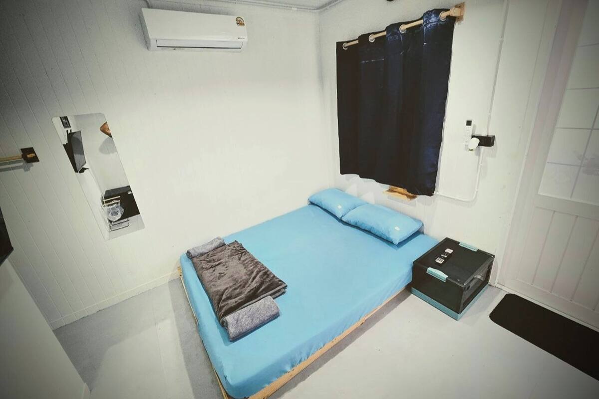 A single bed with blue bedding, a black suitcase, and a window with a black curtain.