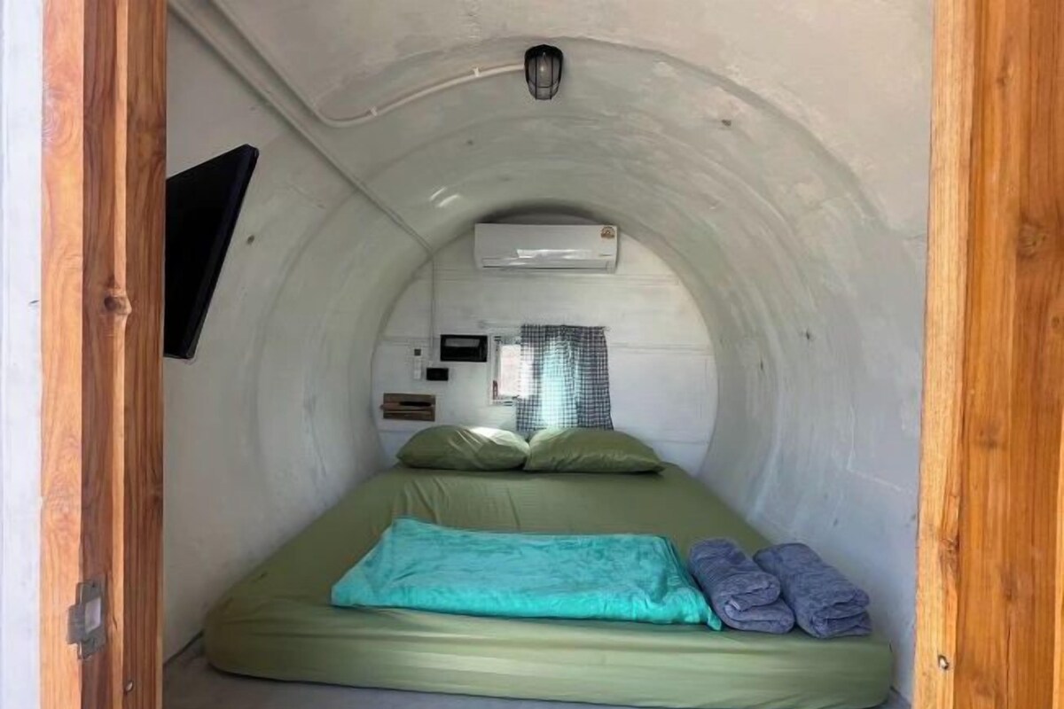 Una habitación pequeña y cerrada con una cama individual, un televisor montado y un equipo de aire acondicionado.