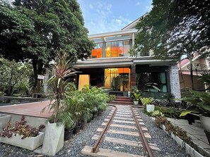 A modern house with a glass facade, surrounded by lush greenery and a gravel pathway. - Onmywayresort (Nakhon Nayok)
