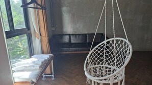 Ein Zimmer mit einem hängenden Makramee-Stuhl, einer Bank mit Kissen, einem schwarzen Ledersofa und einem Fenster mit Vorhängen.