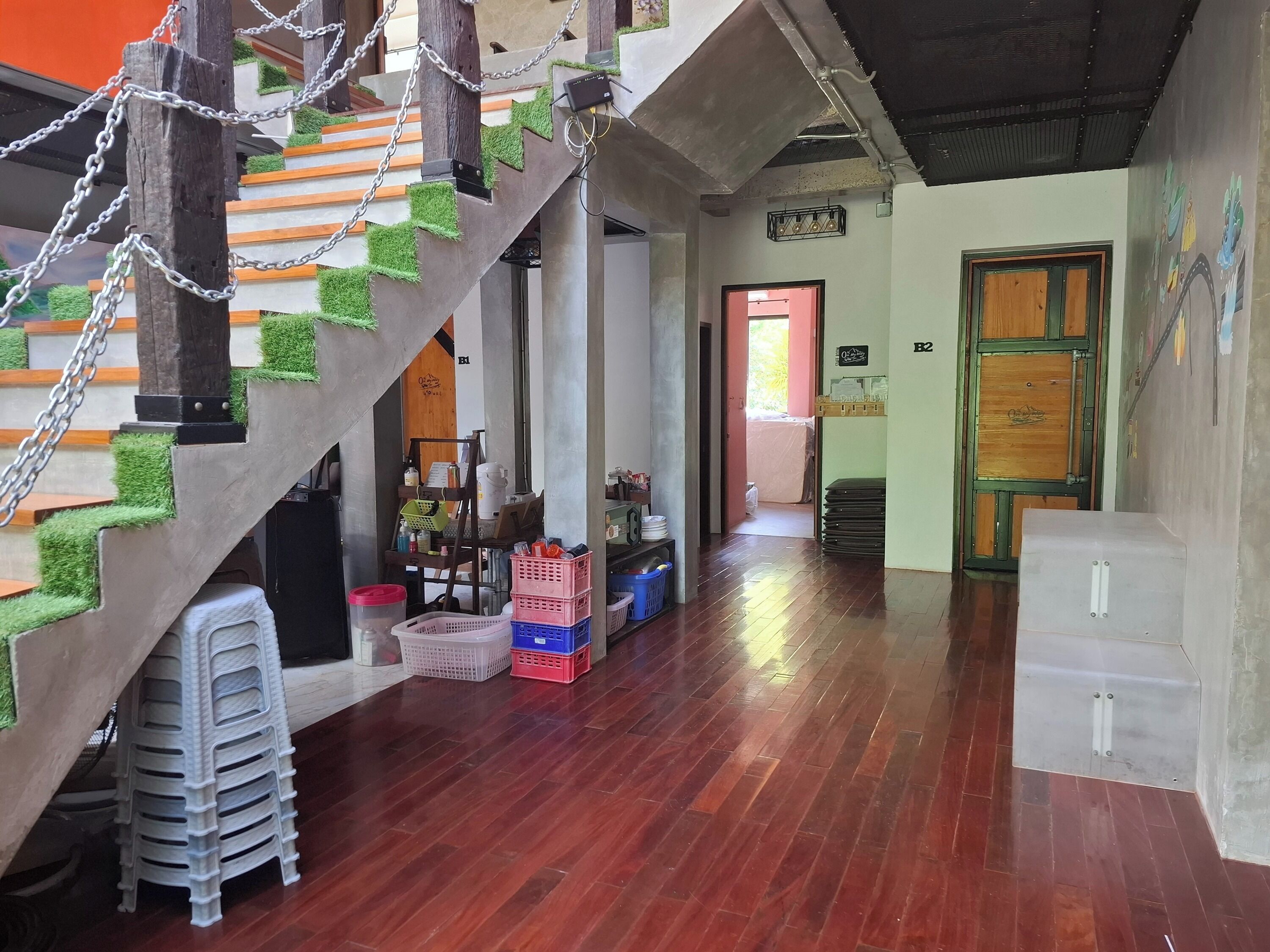 Una escalera con césped artificial verde, escalones de madera y cadenas metálicas. Hay cajas de plástico y un taburete de plástico blanco en el suelo. Se ve una puerta de madera con marco verde al fondo.