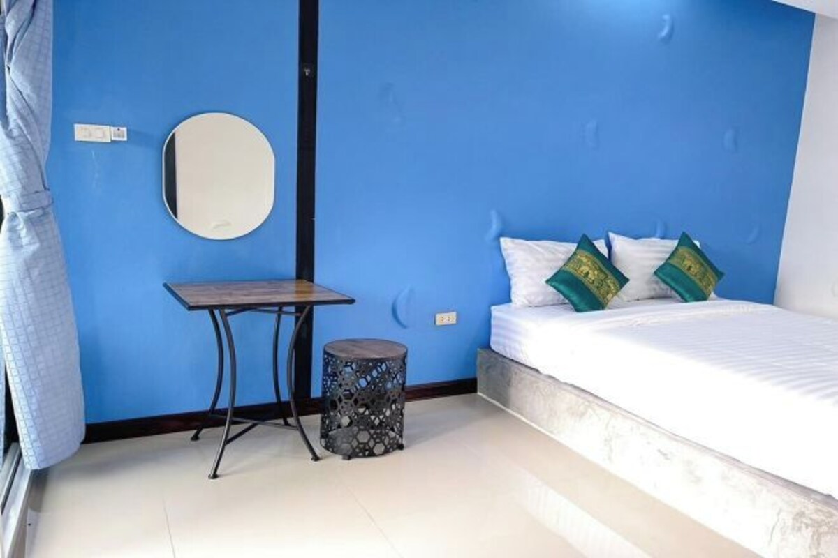 Ein Schlafzimmer mit blauer Wand, einem runden Spiegel, einem kleinen Tisch, einem runden Hocker und einem Bett mit weißer Bettwäsche und grünen Kissen.