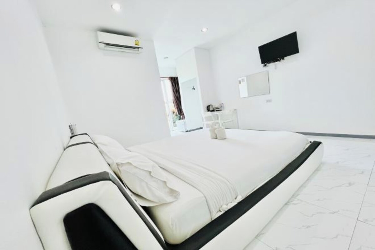 A modern hotel room with a large bed, a flat-screen TV mounted on the wall, and a small bathroom area visible in the background.