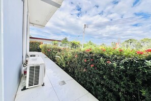 An outdoor area with a white air conditioning unit, a tiled floor, and a hedge with red flowers.