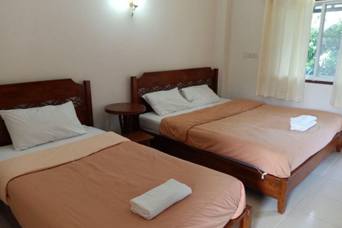A hotel room with two single beds, each with a wooden headboard and a matching brown bedspread. There is a small bedside table with a lamp on it, and a window with curtains.