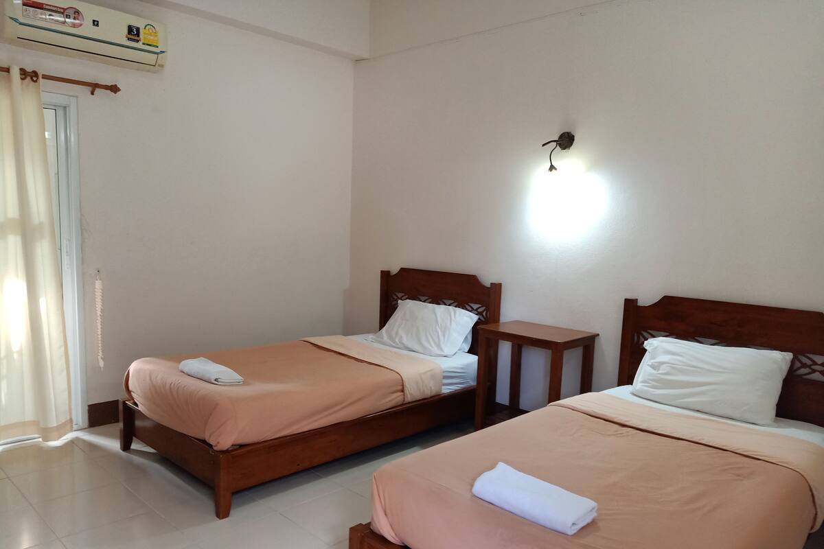 Two single beds with wooden headboards, a small wooden table, and a wall-mounted light fixture in a room with white walls and a tiled floor.