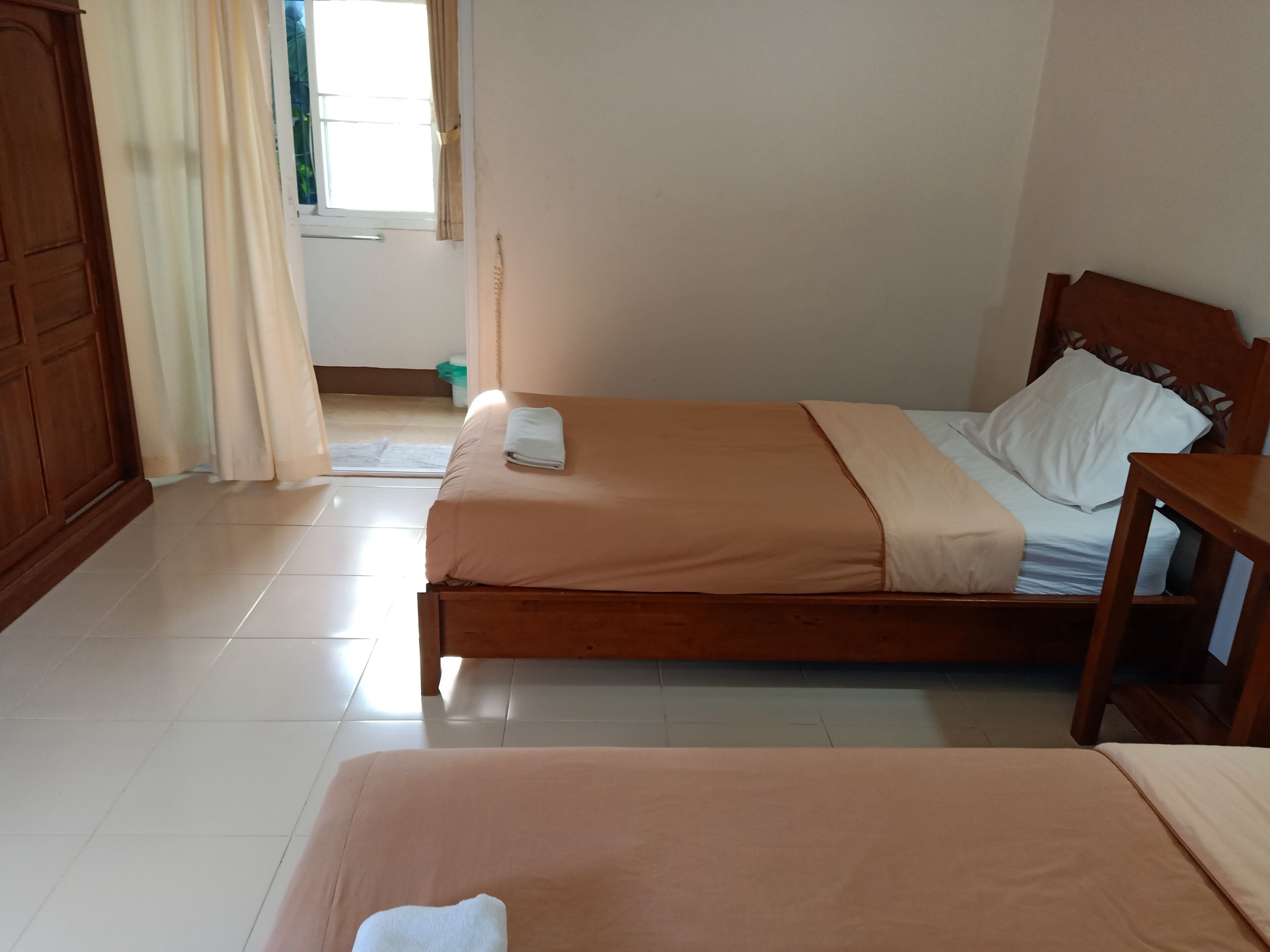 Una habitación con dos camas individuales, cabecera y mesita de noche de madera, una ventana con cortinas y suelo de baldosas.