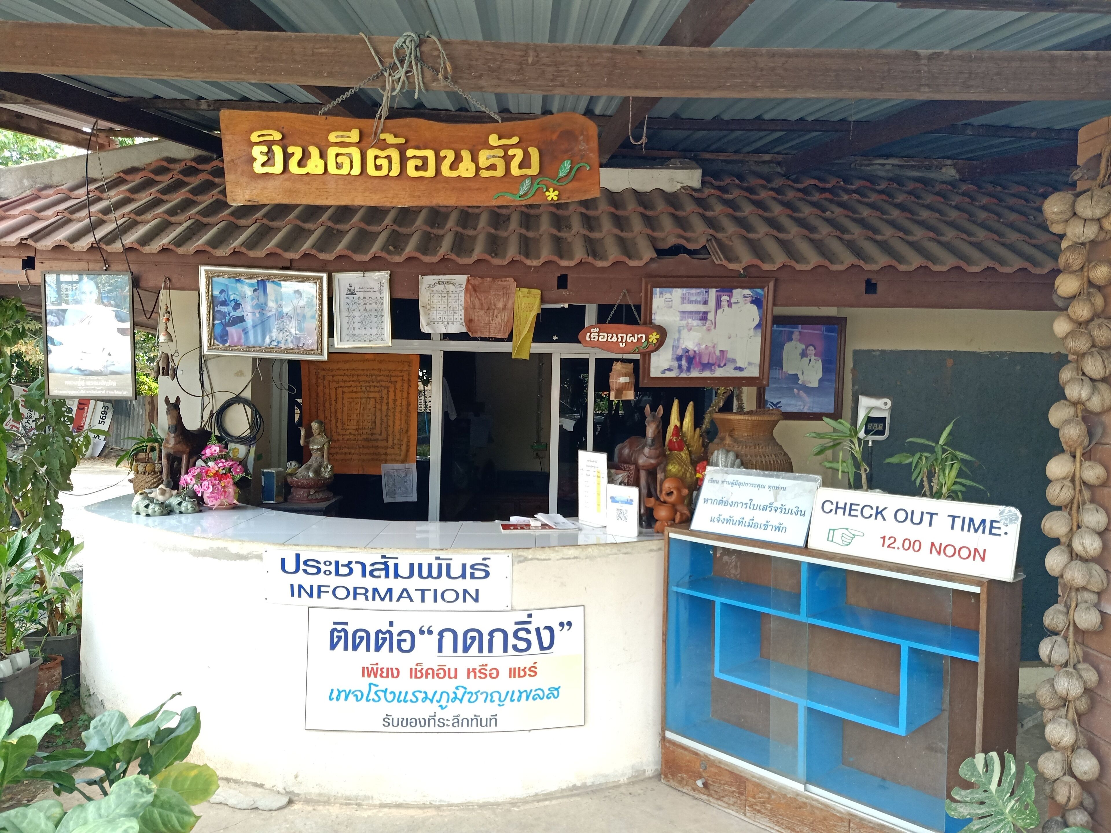 A small, rustic building with a tiled roof and a wooden signboard. There are several framed pictures and a small statue on the wall. A sign indicates a check-out time of 12:00 noon.