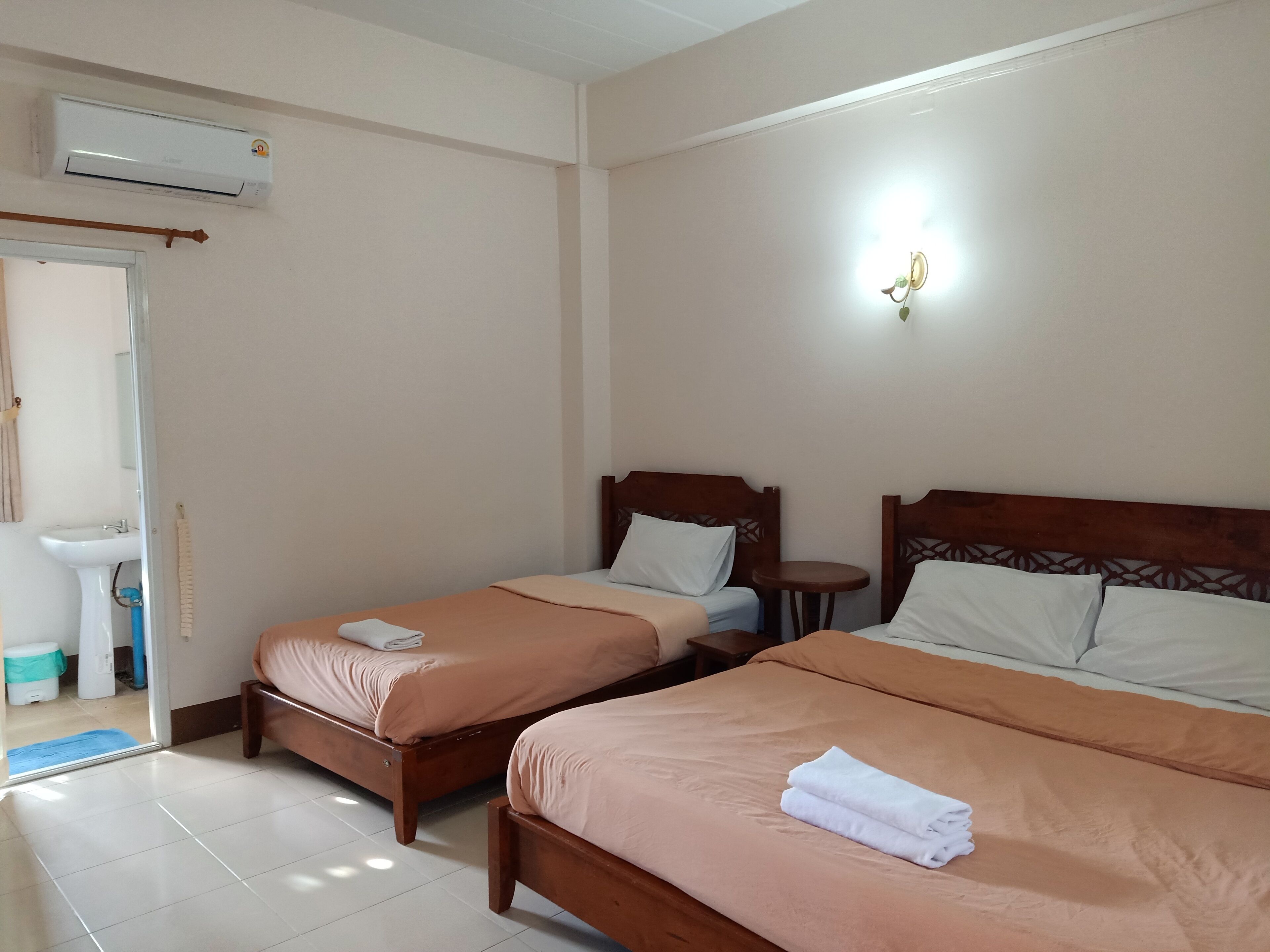 Habitación de hotel con dos camas individuales, una mesita de noche pequeña, aire acondicionado instalado en la pared y un baño visible a través de una puerta abierta.